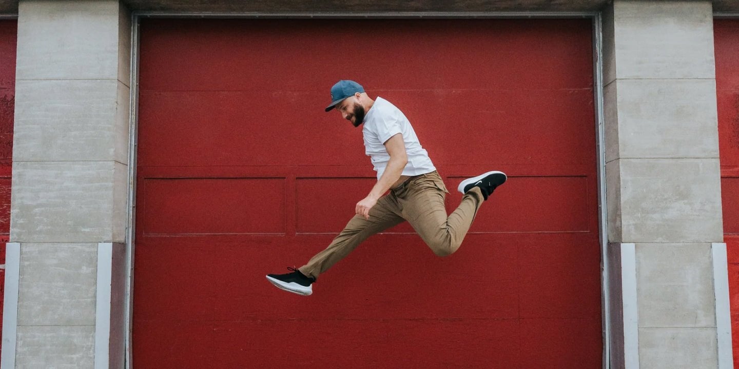 man wearing DUER pants jumping across a red garage door
