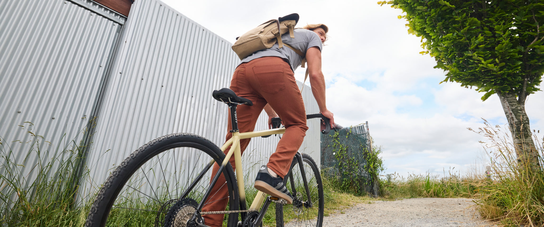 A person riding a bicycle on a gravel path wearing a grey t-shirt, red pants, and a beige backpack. 