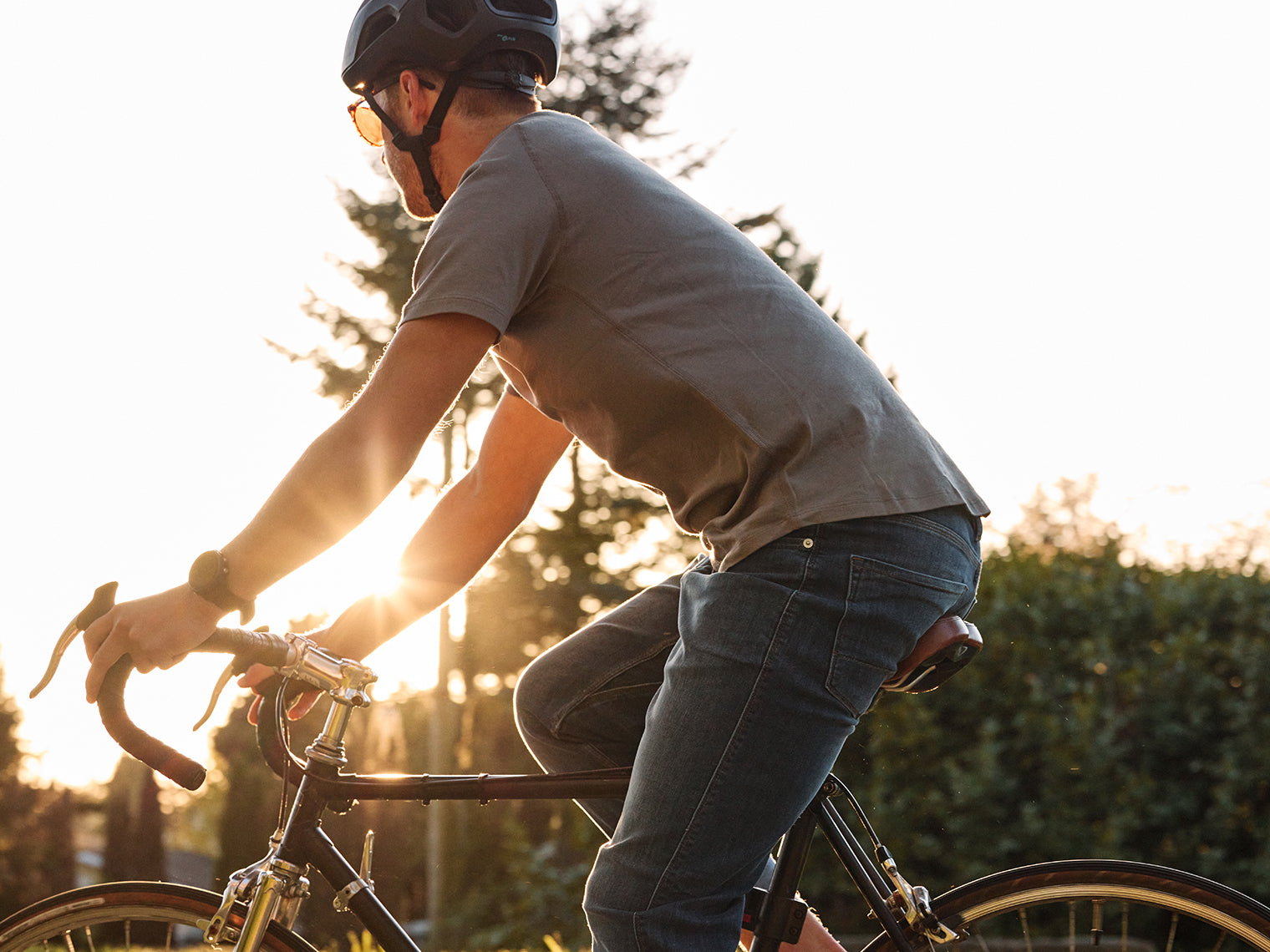 A person wearing a helmet and sunglasses rides a bicycle outdoors during sunset. They are dressed in a grey T-shirt and jeans