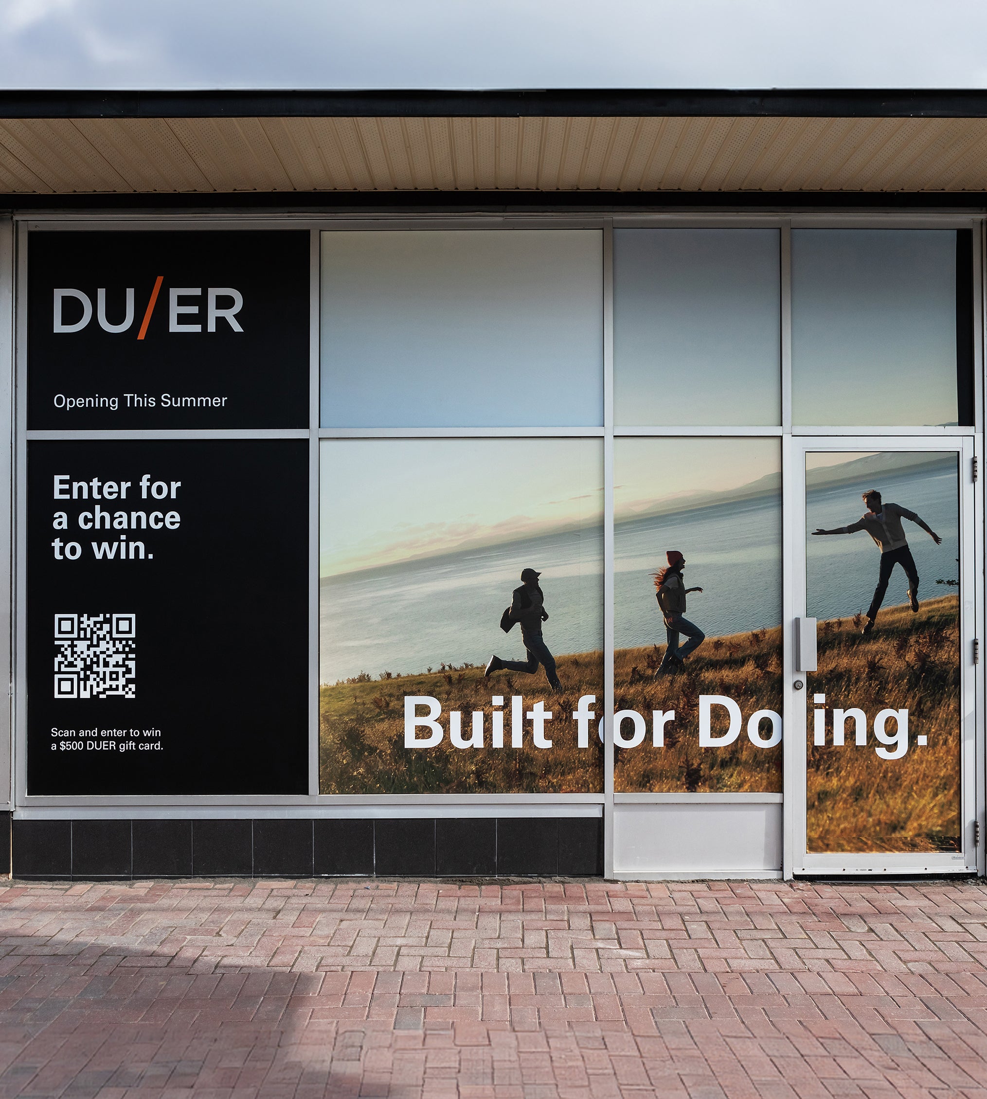 DUER storefront with large windows displaying an ad of people running on a hill by the water. Text reads 'Built for Doing.' QR code and 'Enter for a chance to win.' are also visible, along with 'Opening This Summer'.