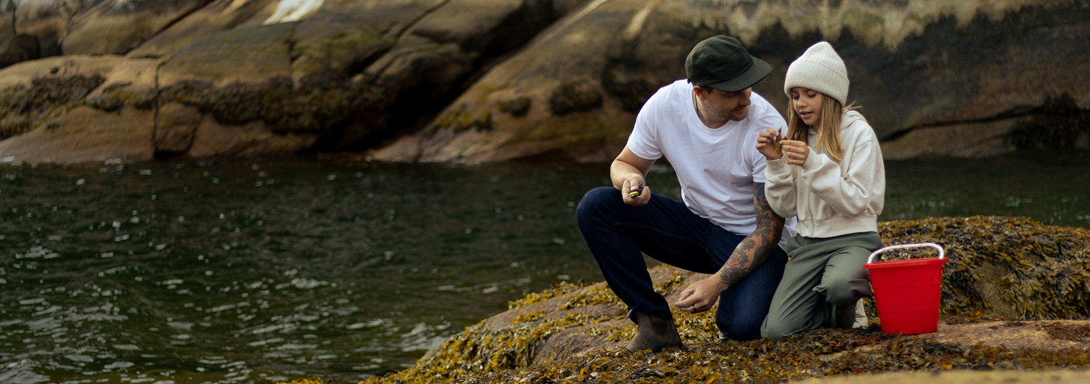 A man and a girl are crouching on a rocky shore by the water, examining items in their hands. The man wears a white t-shirt, jeans, and a cap, while the girl is dressed in a beanie, jacket, and pants. 