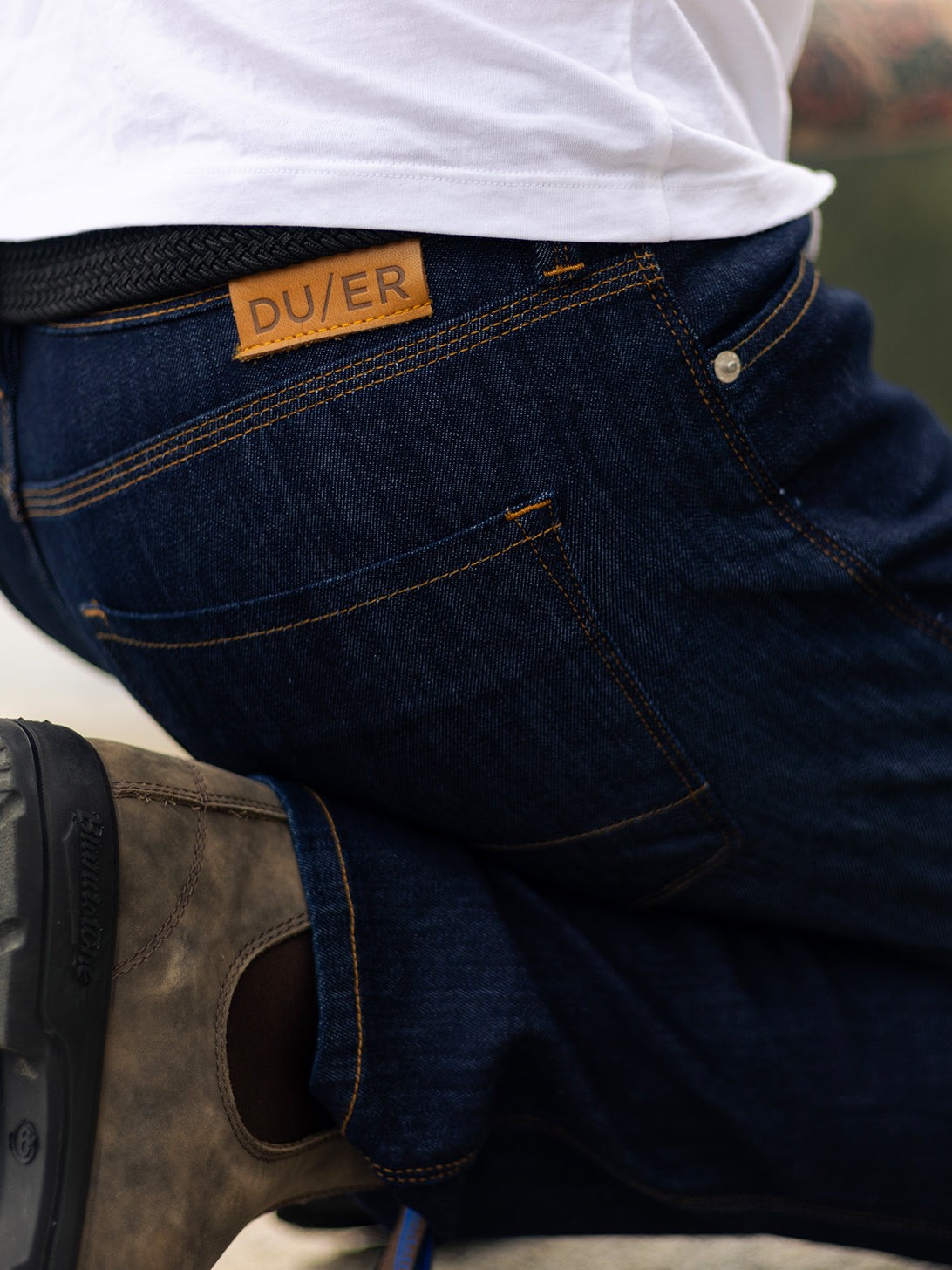 Man kneeling down wearing a white shirt and dark jeans with a brown leather patch labeled "DU/ER" on the back pocket is seen from behind. 