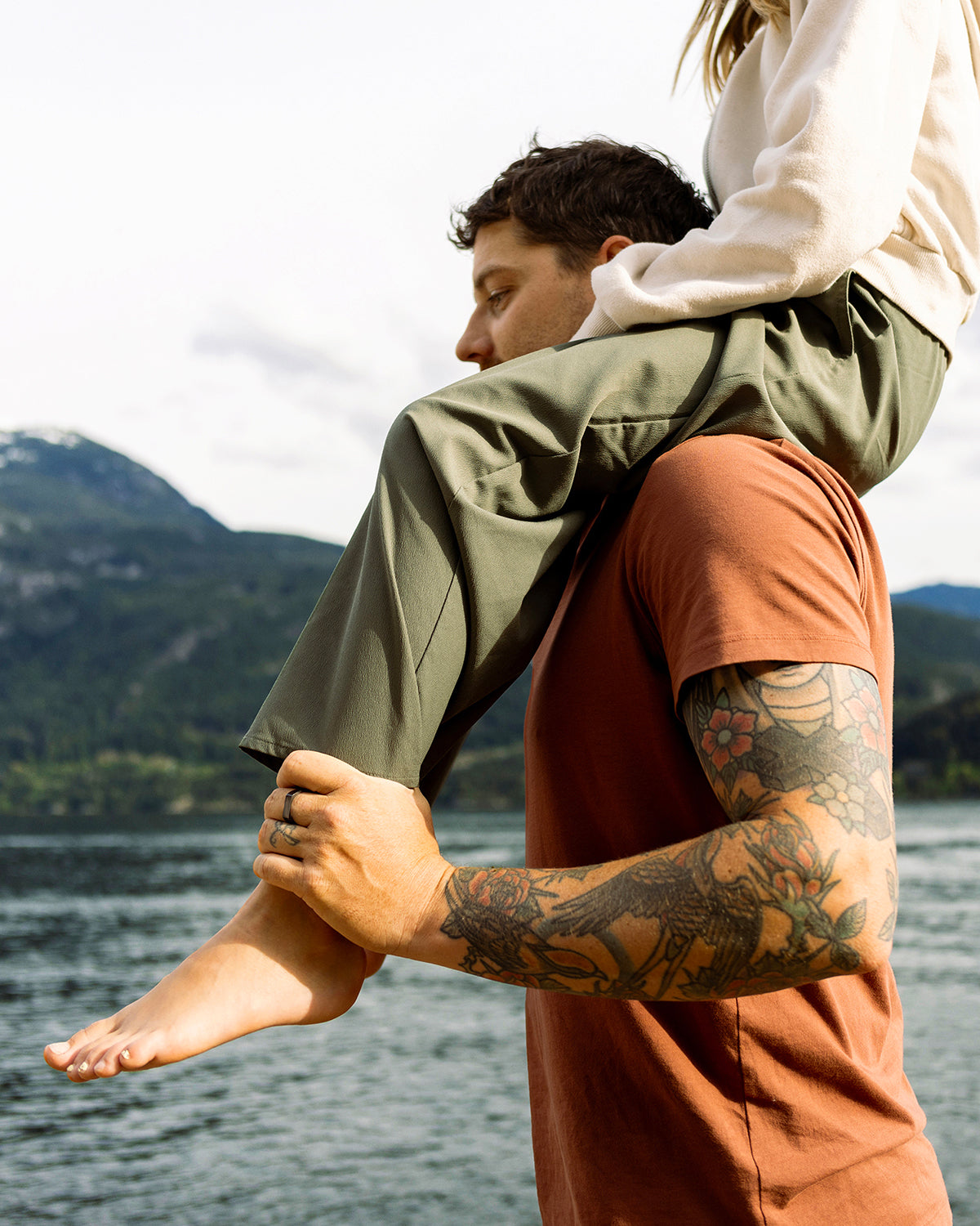 A man with tattoos carries a woman on his shoulders by a large scenic lake.
