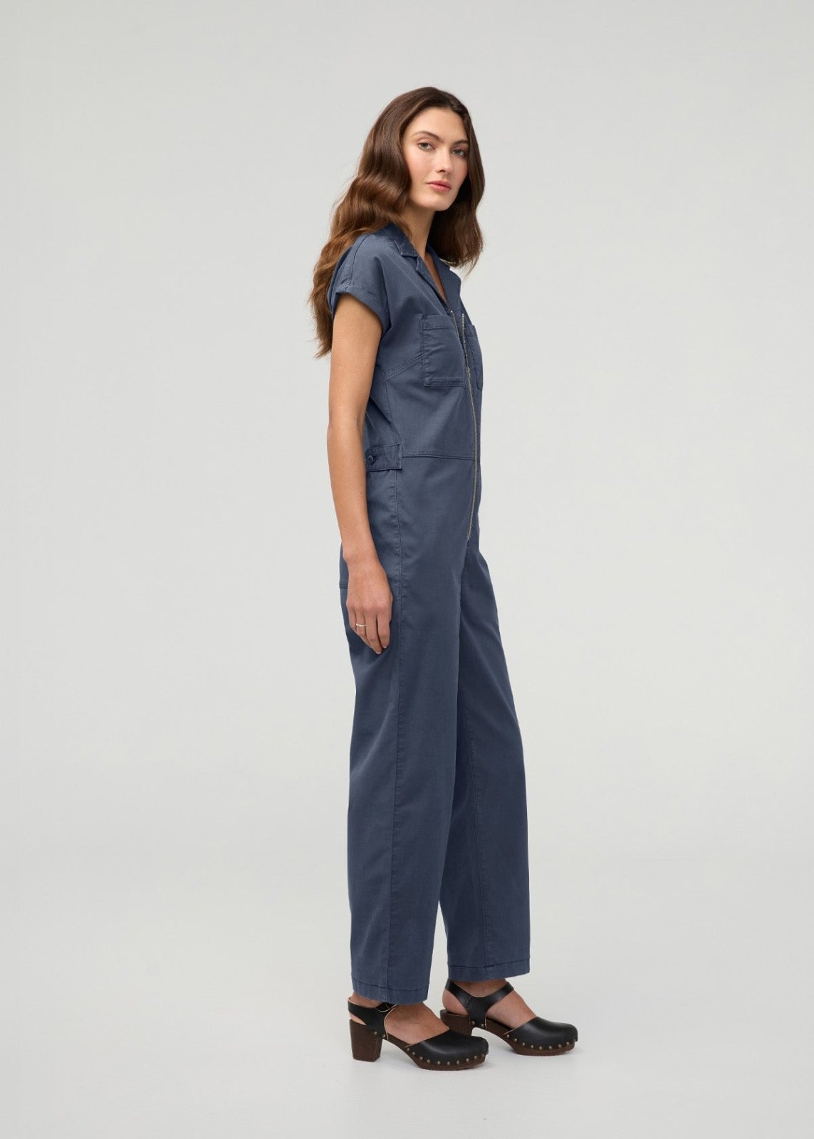 Woman wearing a blue boiler suit with short sleeves, front zipper, and chest pockets, paired with black platform clogs.