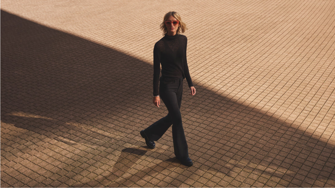 A woman with red sunglasses walks on tiled pavement, wearing a black long-sleeved top and black pants.