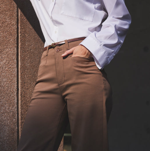 Person standing against a wall, wearing brown trousers and a white blouse