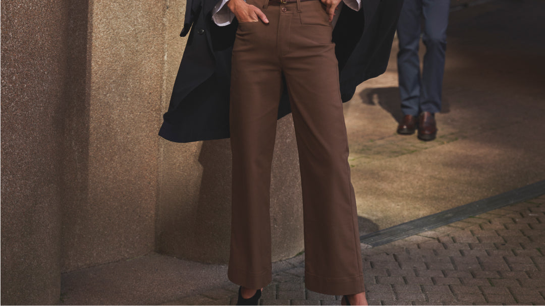 A woman standing on a paved sidewalk wearing brown pants, black heels, and a black coat