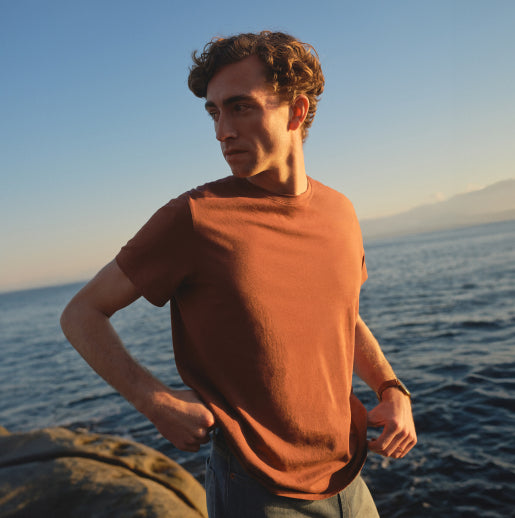 A man wearing a brown t-shirt and jeans stands on a rocky shoreline, gazing into the distance as the sun sets over the ocean behind them. 